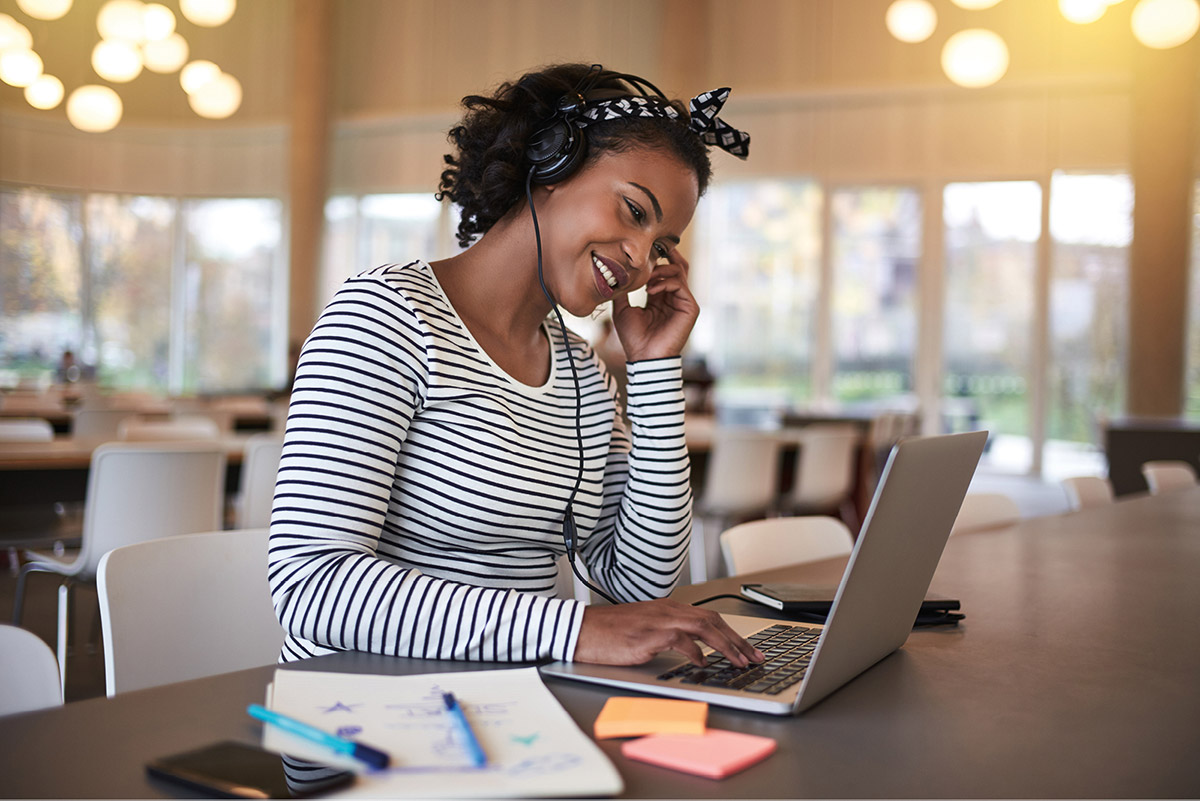 a student in a striped shirt wearing headphones and smiling while studying on a computer