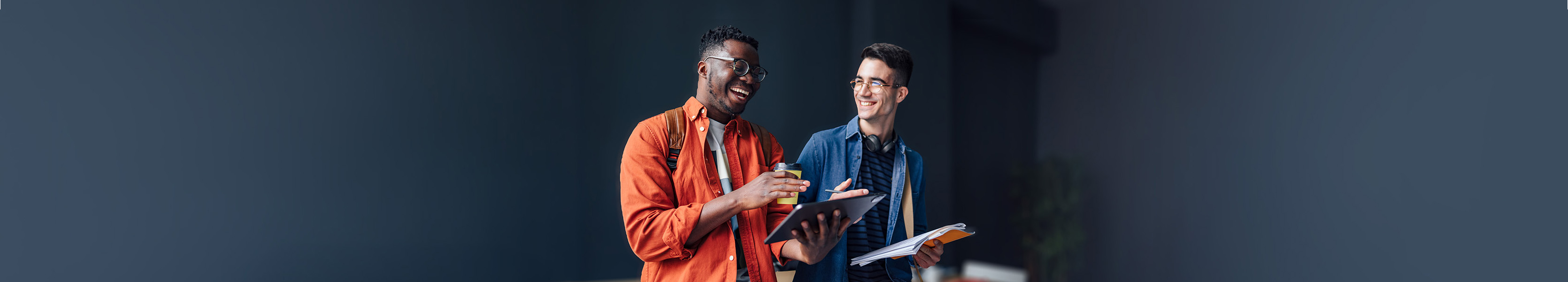 Smiling student holding a digital tablet and cup of coffee while standing and laughing with his friend.